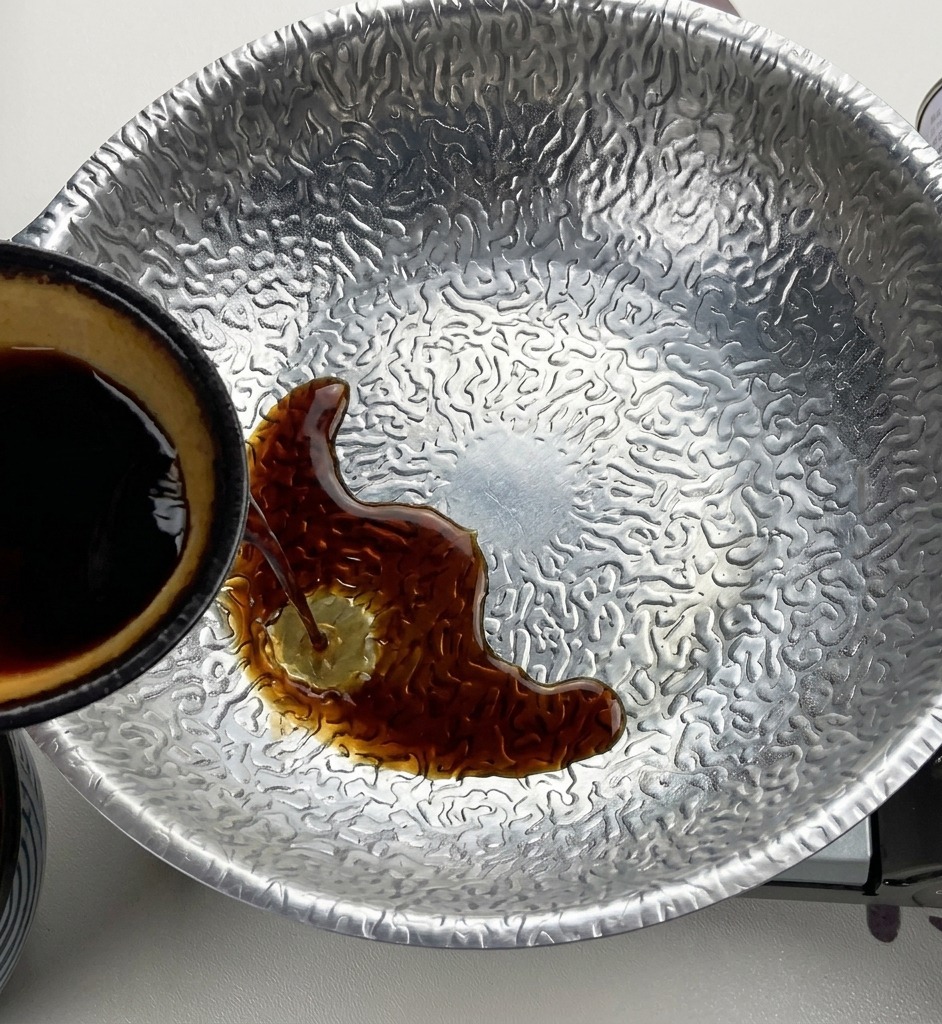 Dark soy sauce being poured from a small bowl with a yellow interior into a textured metal pot containing miso and sugar.