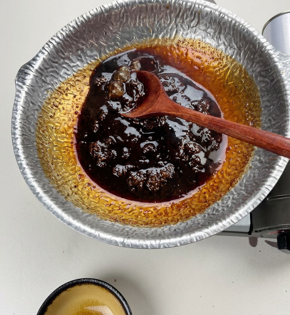 A wooden spoon stirring a thick, dark brown, bubbling sauce in a hammered silver-colored pot.