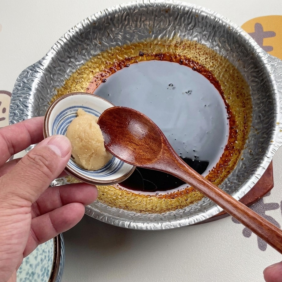 A wooden spoon holding a scoop of light-colored white miso paste over a dark, simmered sauce in a metal pot.