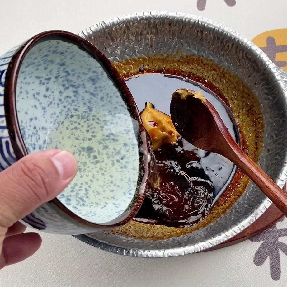 Clear dashi broth being poured from a blue and white patterned ceramic bowl into the dark miso sauce.