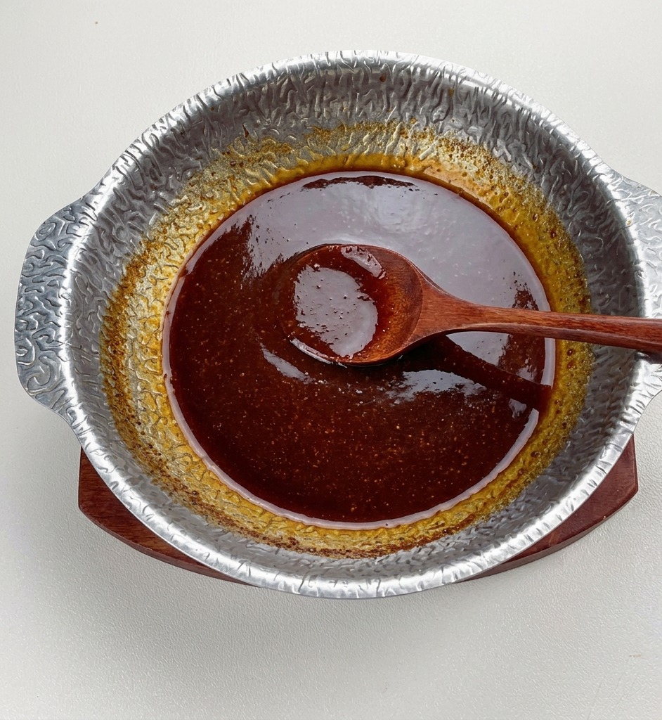 The finished dark miso sauce simmering in a metal pot, appearing thick and shiny with a wooden spoon resting in the center.