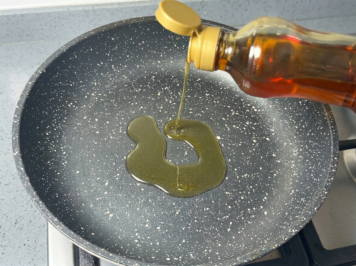 Cooking oil being poured from a bottle into a dark, speckled non-stick frying pan.