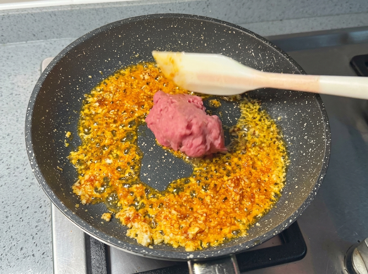 A portion of raw minced pink pork being placed into a pan of sizzling aromatics with a white silicone spatula.