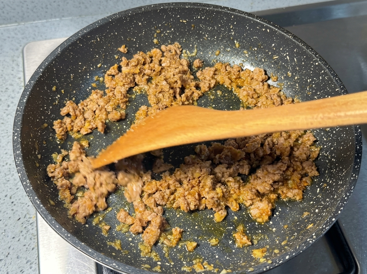 Browned minced pork being stir-fried in a speckled pan with a wooden spatula.