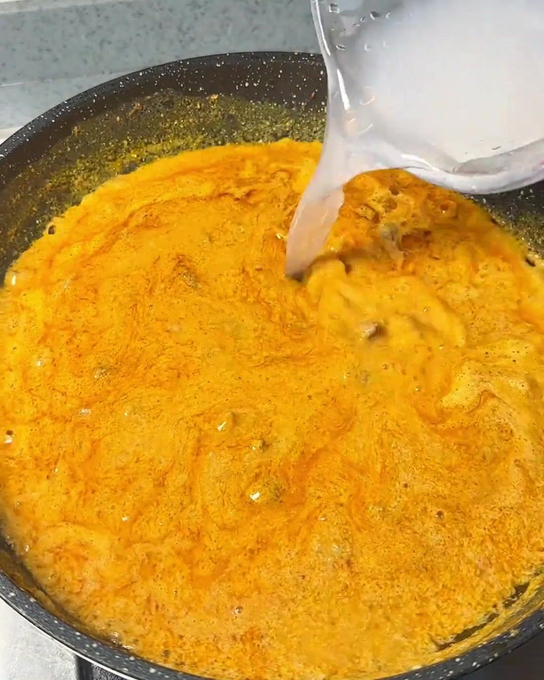Clear water being poured from a container into a bubbling, rich orange curry mixture in a dark pan.