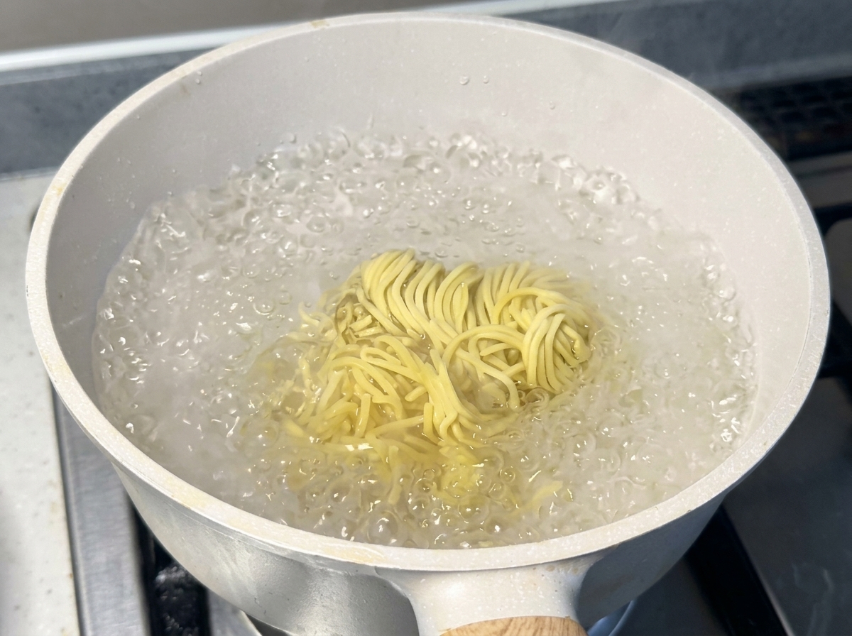 A cluster of fresh yellow ramen noodles boiling vigorously in a white pot filled with bubbling hot water.