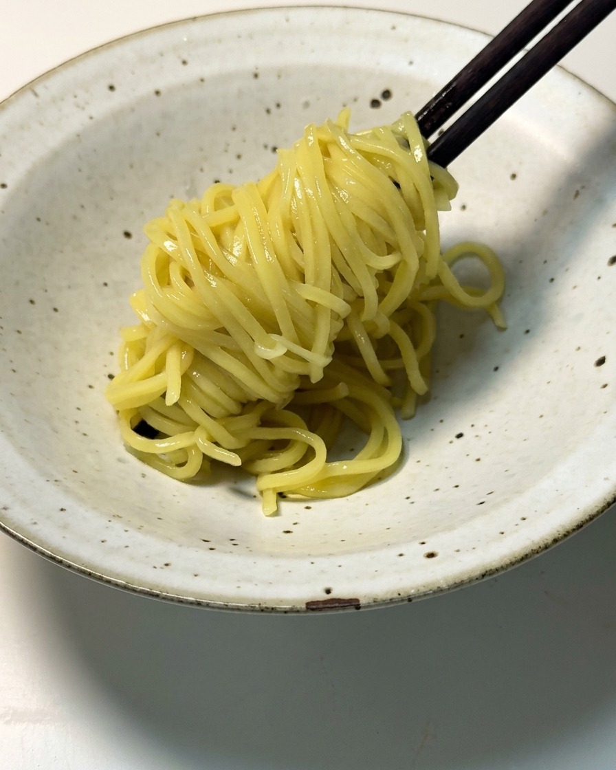 Cooked yellow ramen noodles being lifted with dark wooden chopsticks and placed into a shallow, speckled ceramic bowl.