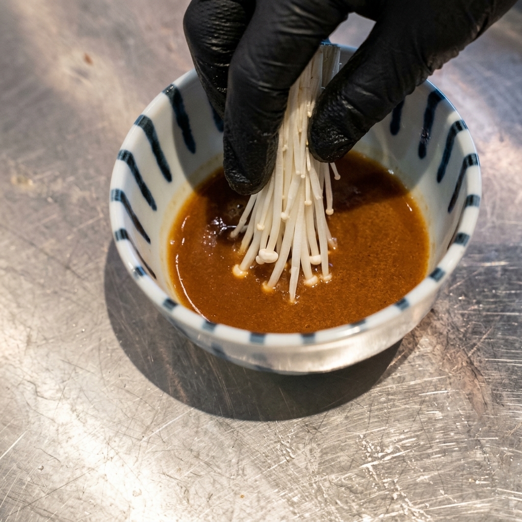 A hand wearing a black glove placing a cluster of white bean sprouts into a bowl of brown dipping sauce.