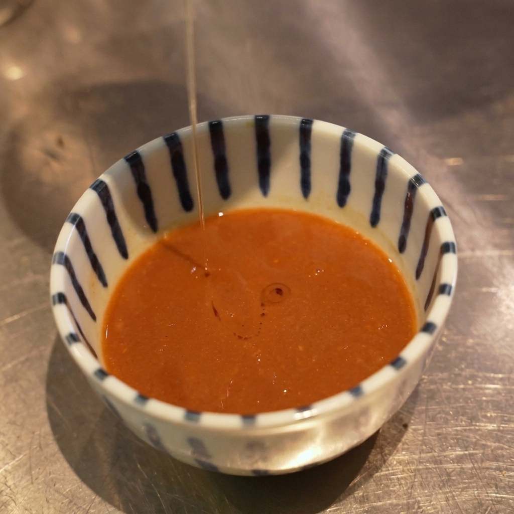 A thin stream of orange-tinted oil being drizzled from above into a bowl of dipping sauce, forming droplets on the surface.