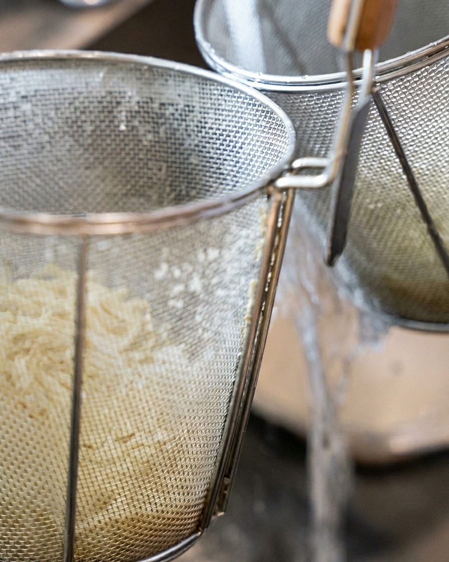 Thick ramen noodles inside a cylindrical mesh strainer being rinsed under a stream of water.