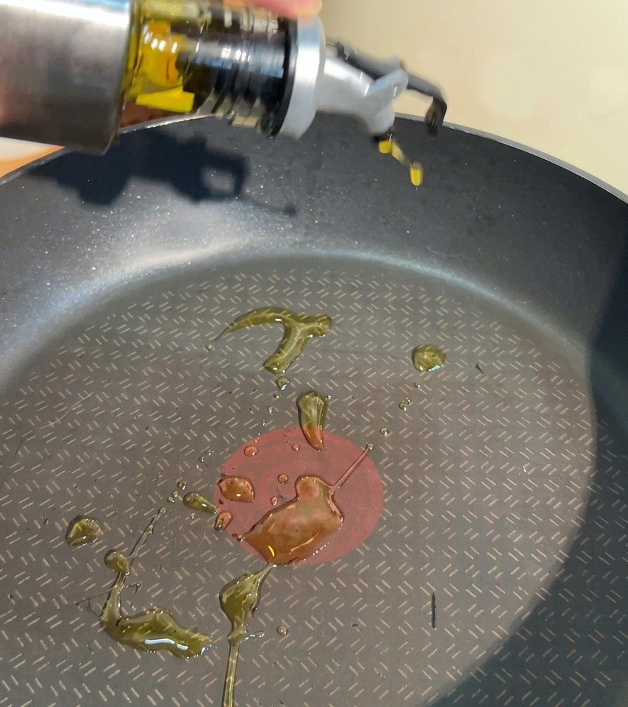 A hand pouring clear cooking oil from a stainless steel dispenser into a black non-stick frying pan.