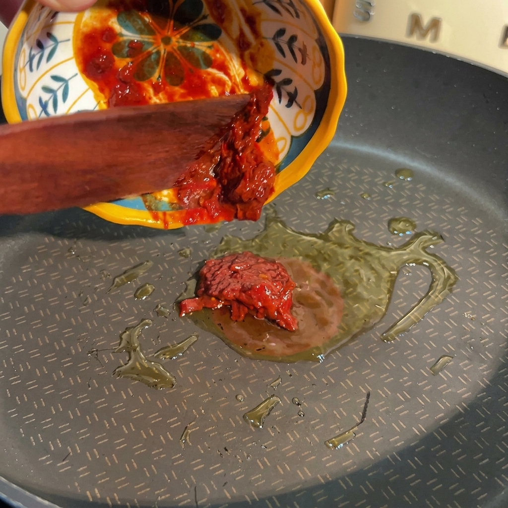 A wooden spatula pushing thick red bean paste from a decorative yellow and blue bowl into a pan with hot oil.