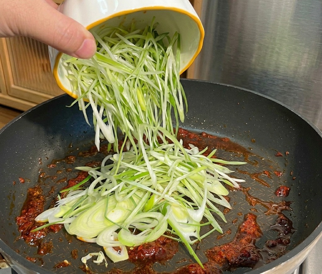 Finely sliced green onions being poured from a white bowl with a yellow rim into a frying pan containing red sauce.
