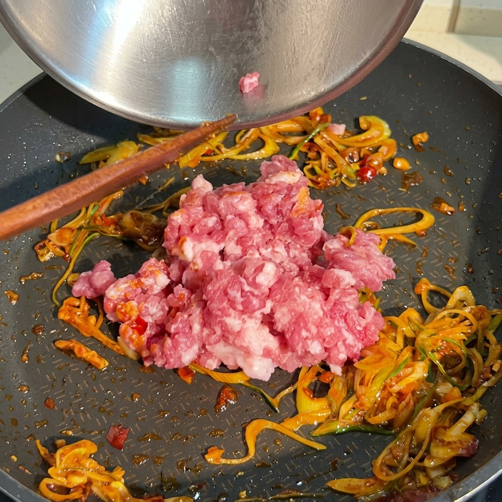 A pile of raw pink minced pork being added to a frying pan already containing sautéed green onions and red chili sauce.