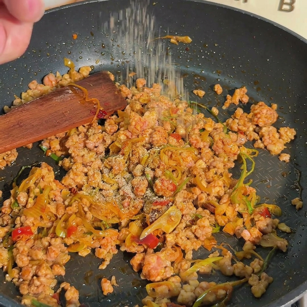 A close-up shot of white pepper being sprinkled over browned minced pork and green onions in a black frying pan.