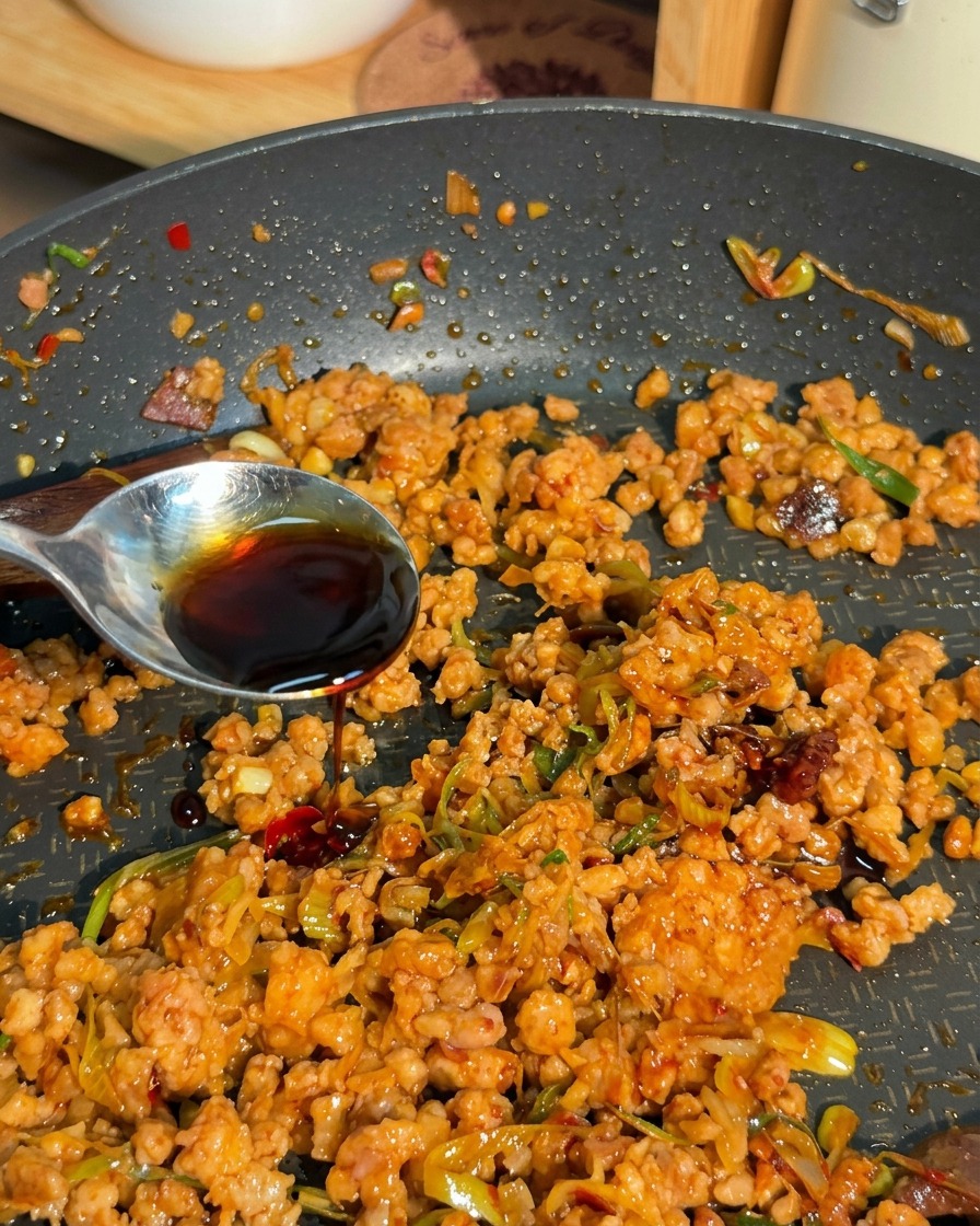 Dark savory liquid being poured from a silver spoon over a mixture of browned minced meat and green onions in a pan.