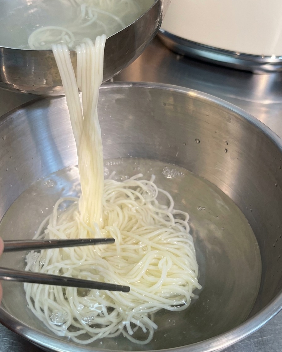 Silver chopsticks lifting a large portion of cooked white noodles from a metal bowl of water into another bowl.
