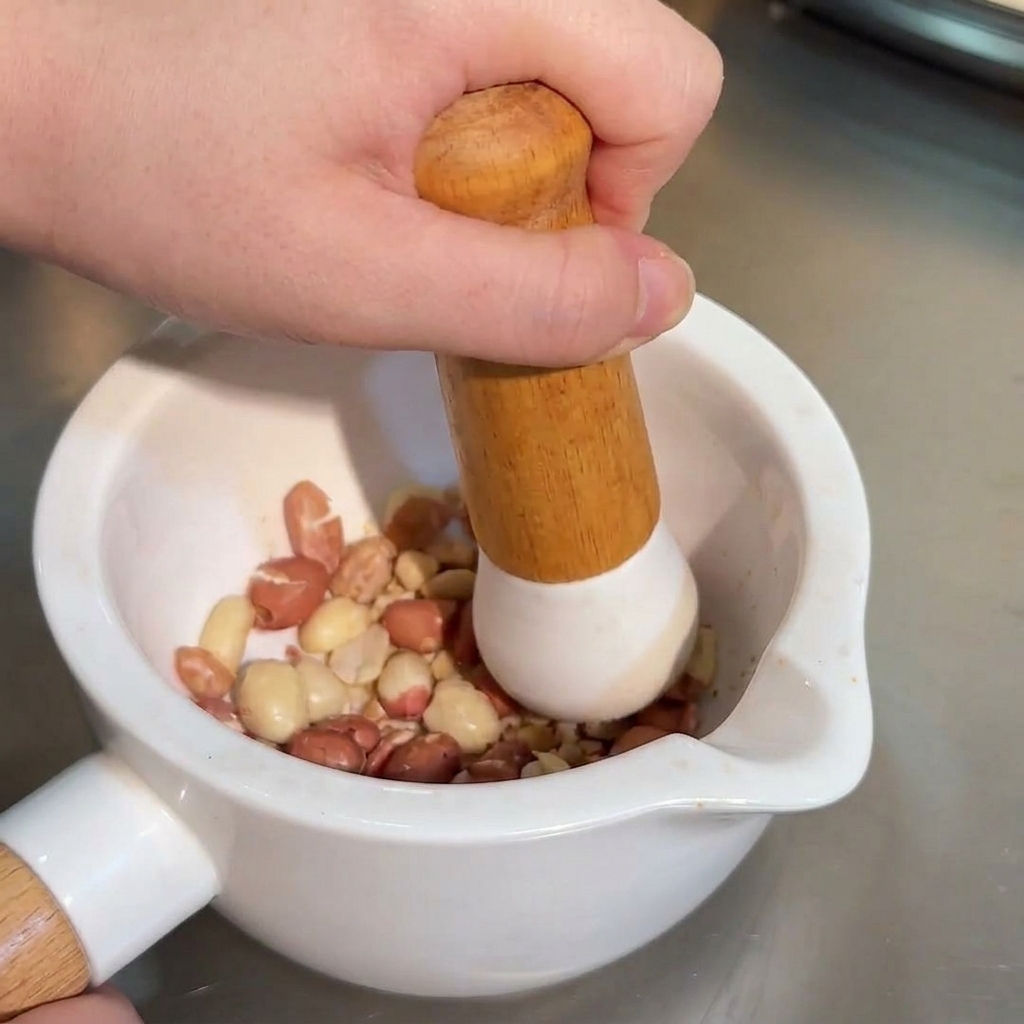 A hand using a wooden pestle to crush whole roasted peanuts inside a white ceramic mortar.