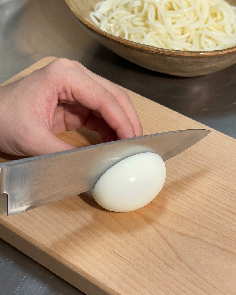 A hand holding a knife to slice a white soft-boiled egg on a wooden cutting board, with a bowl of noodles in the background.