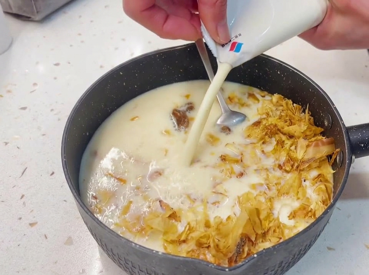 Hands pouring creamy white soy milk from a carton into a dark saucepan containing a mixture of tuna and dried bonito flakes.