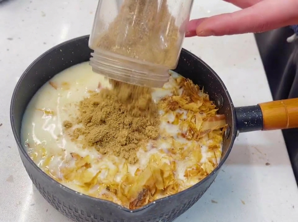 A plastic blender cup pouring finely ground roasted sesame seeds into a pot filled with a milky white liquid, tuna, and bonito flakes.