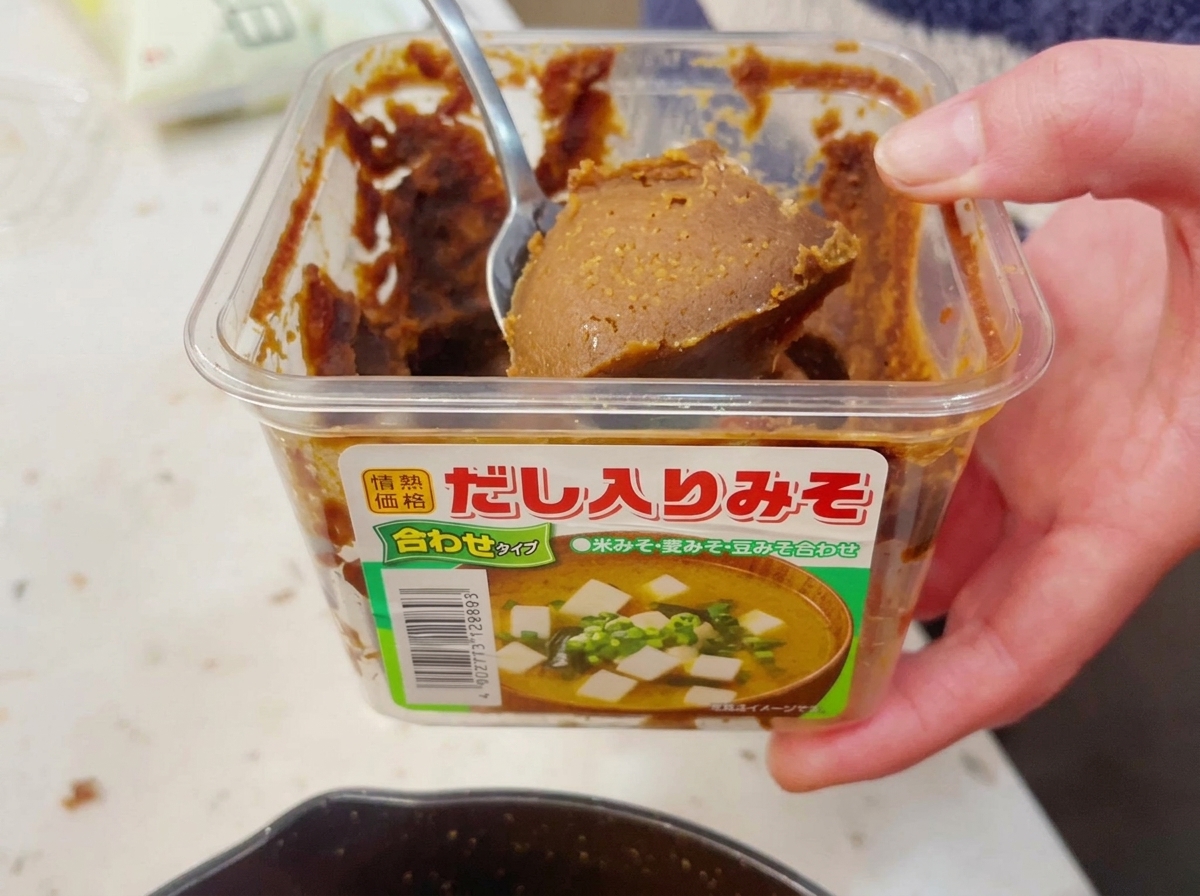A hand scooping a generous spoonful of thick brown miso paste out of a clear plastic container with a metal spoon.