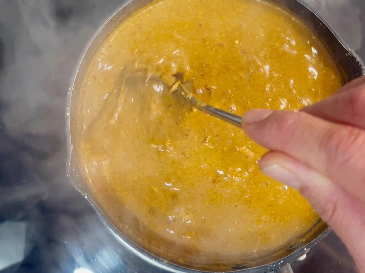 A hand using a metal spoon to stir a thick, bubbling orange-brown dipping broth in a small pot.