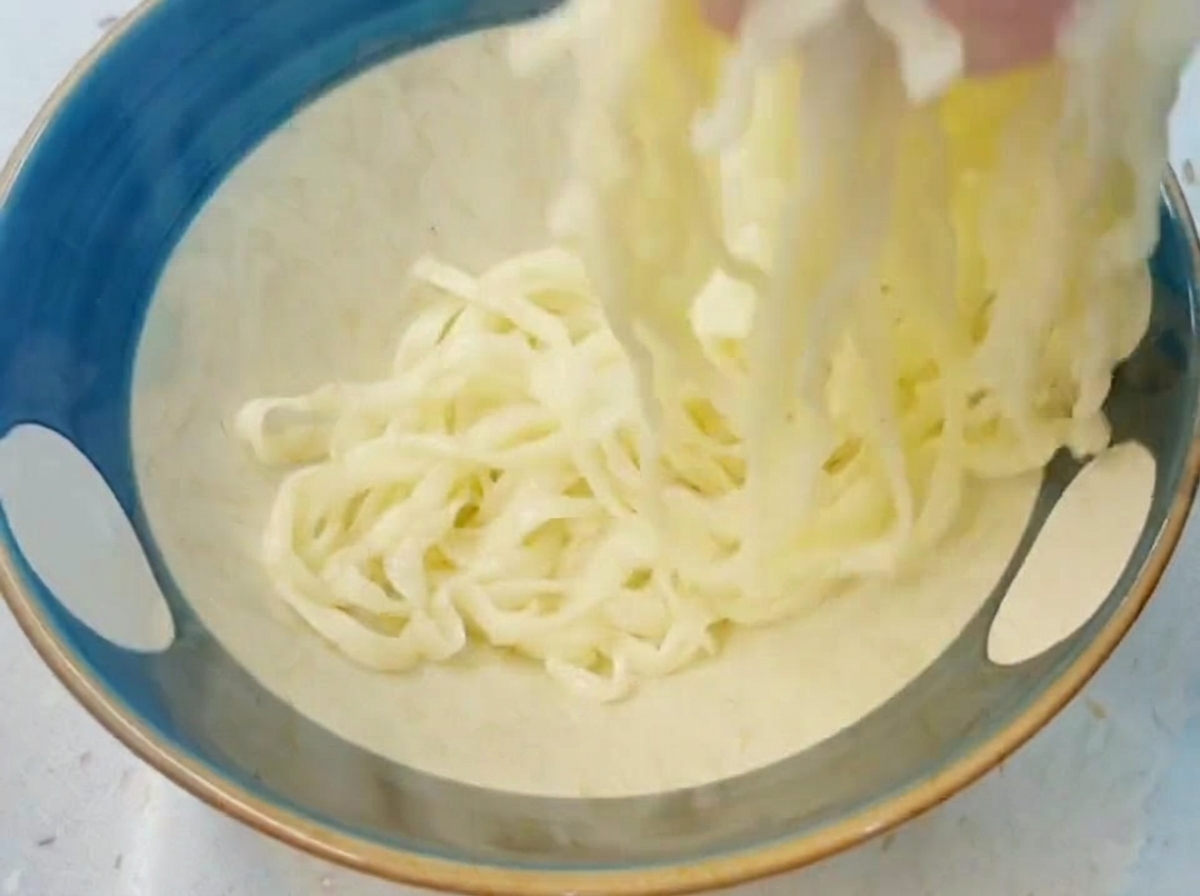 A hand lifting and tossing thick, cooked white ramen noodles inside a blue bowl filled with cold water.