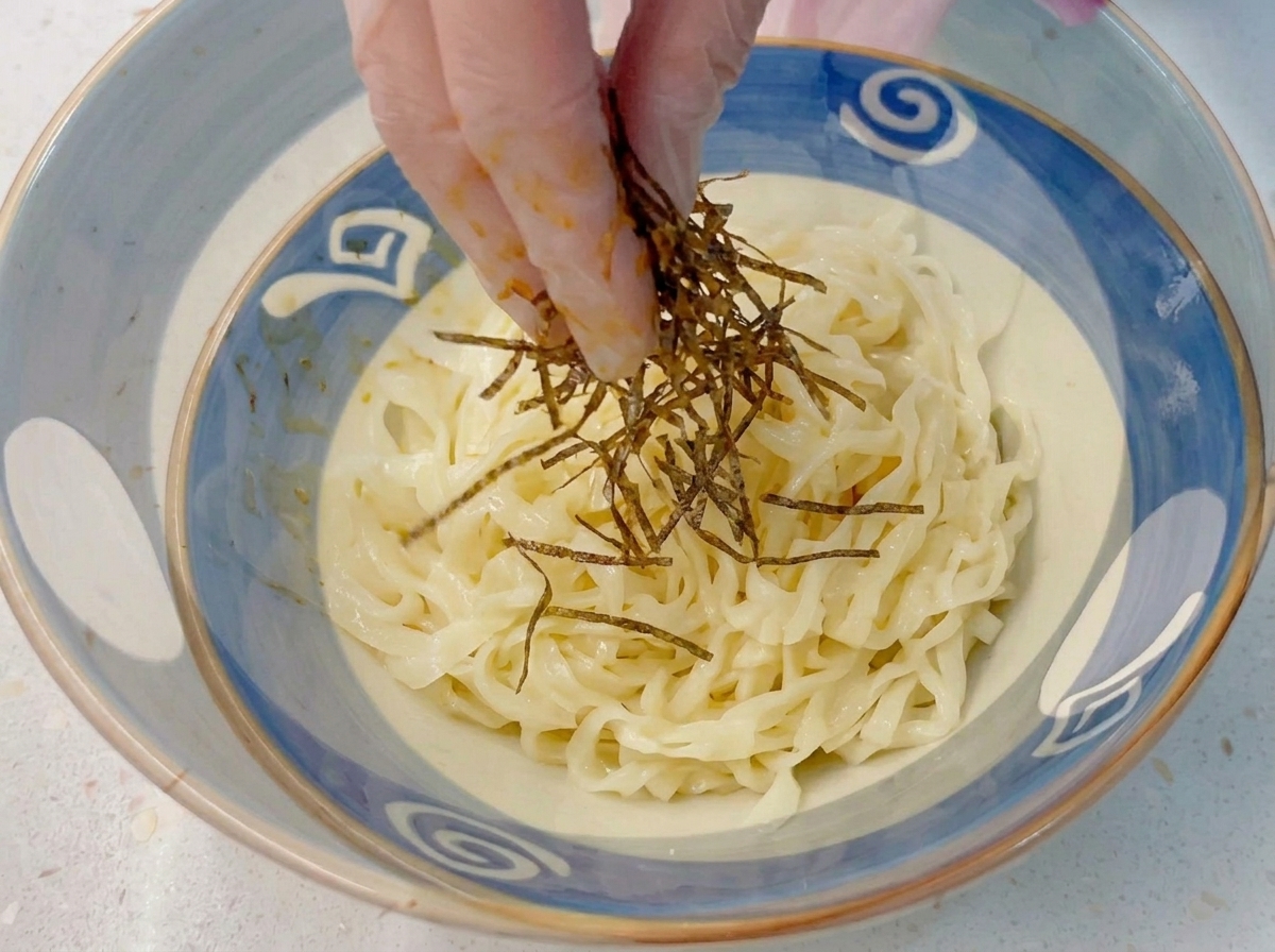 Gloved fingers dropping shredded nori seaweed onto a bowl of cold, thick ramen noodles.