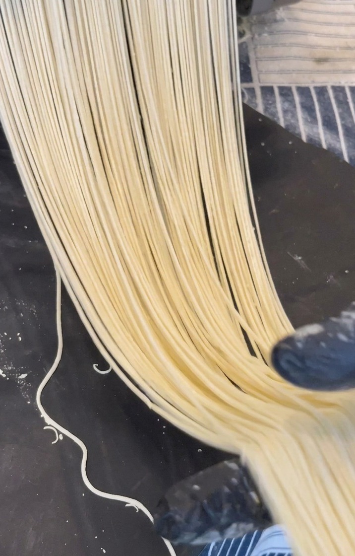 Long strands of pale yellow ramen noodles being extruded from a metal machine onto a dark surface.