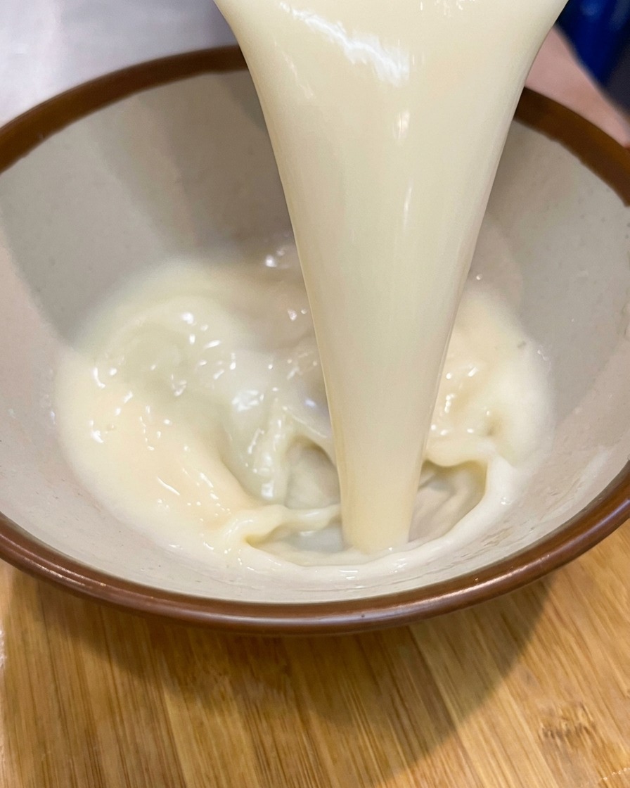 A thick stream of creamy, pale soup base being poured into a brown-rimmed ceramic bowl.