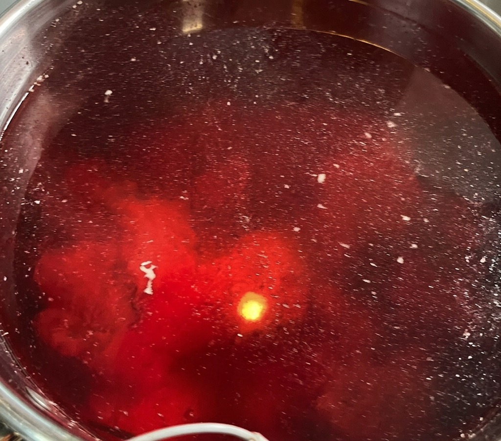 A top-down view of a large stainless steel pot where pork bones are being blanched, with reddish-tinted water and white foam impurities visible on the surface.