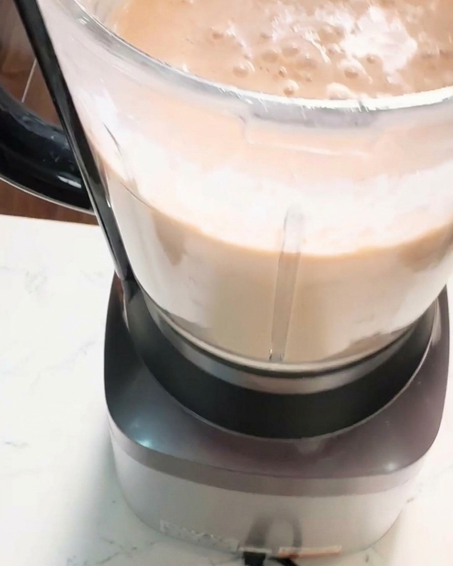 A close-up shot of a blender jar filled with a thick, smooth, creamy orange-pink shrimp paste being processed.