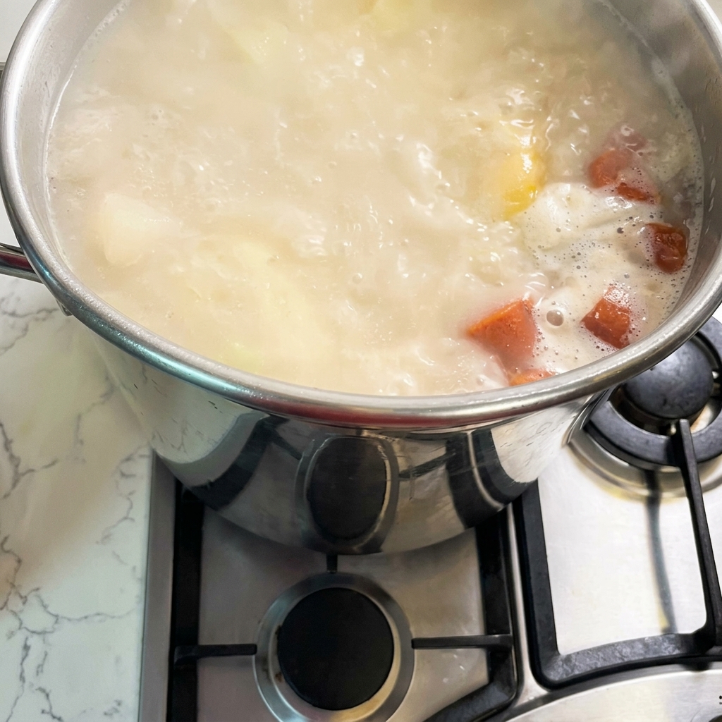 A large stainless steel stockpot on a gas stove filled with bubbling, creamy white and orange broth.