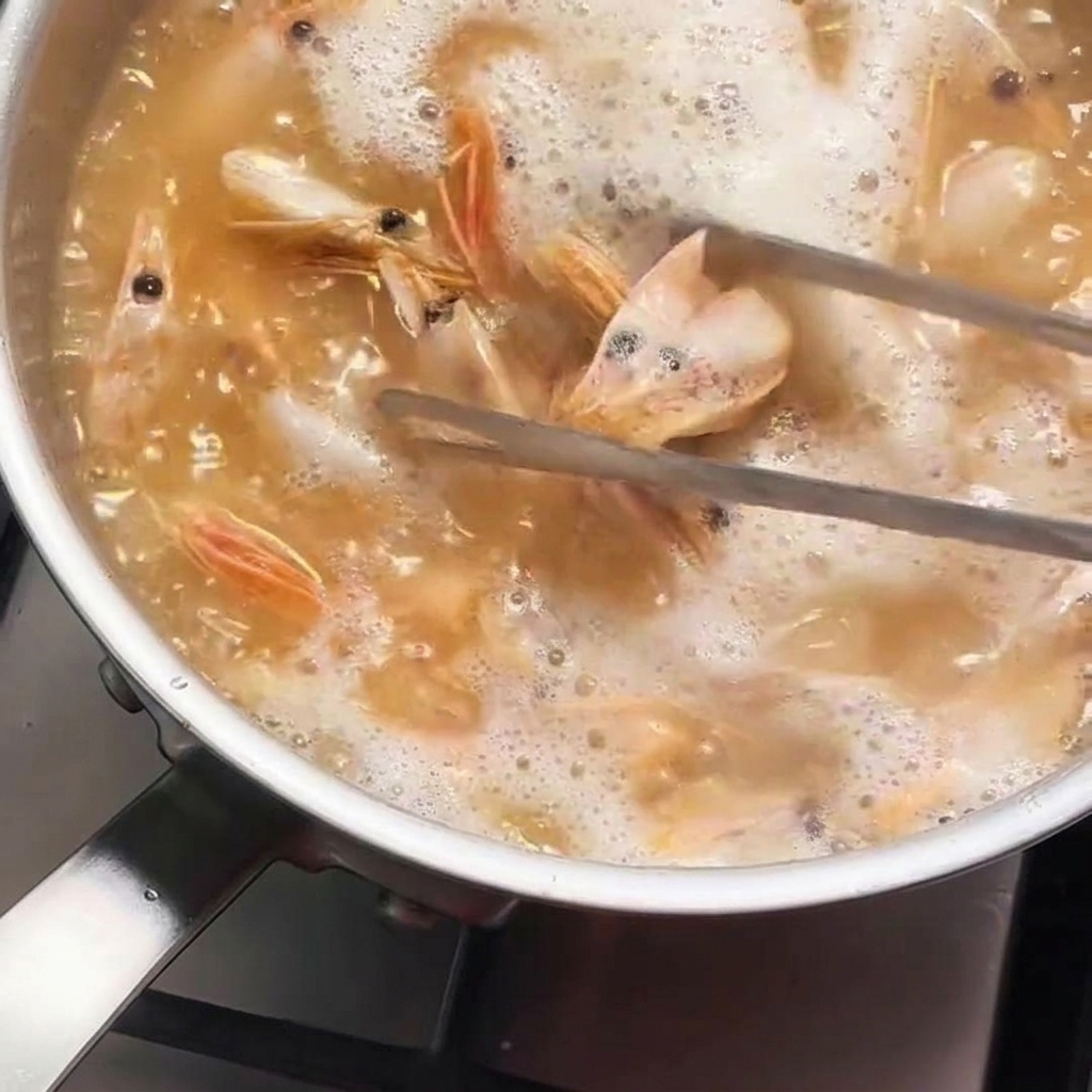 Shrimp heads being stirred with metal tongs in a pot of hot, bubbling oil that is turning a deep orange color.
