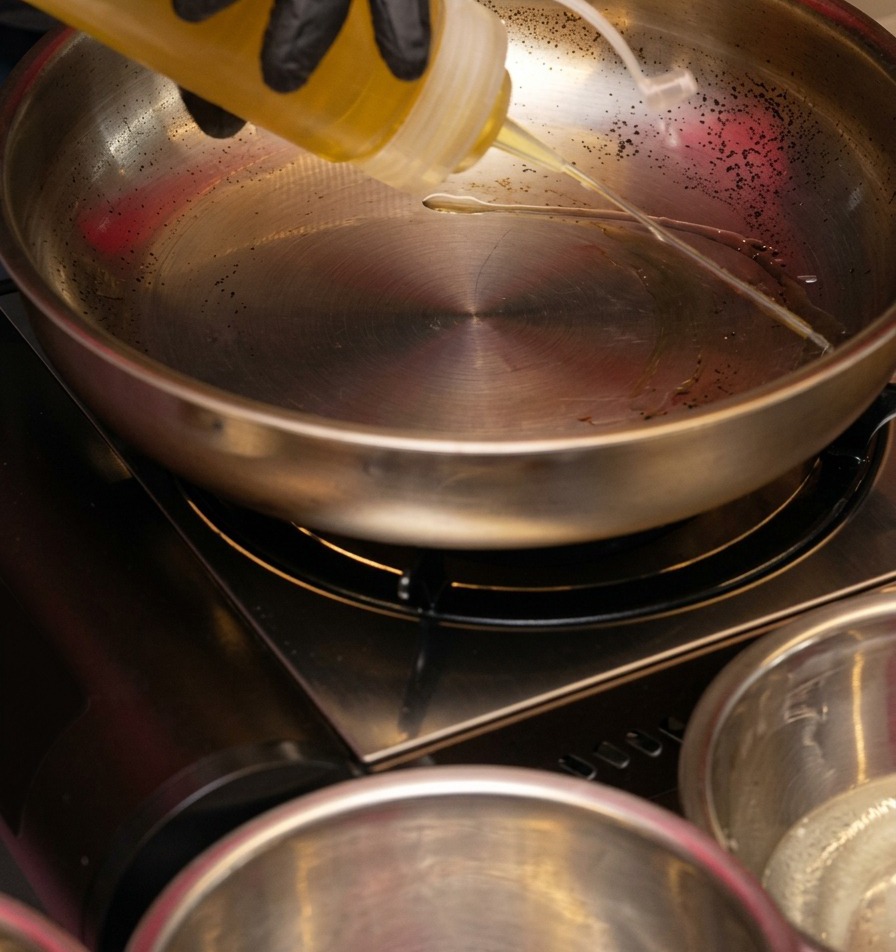 Clear cooking oil being poured from a plastic squeeze bottle into a gleaming stainless steel frying pan on a portable burner.