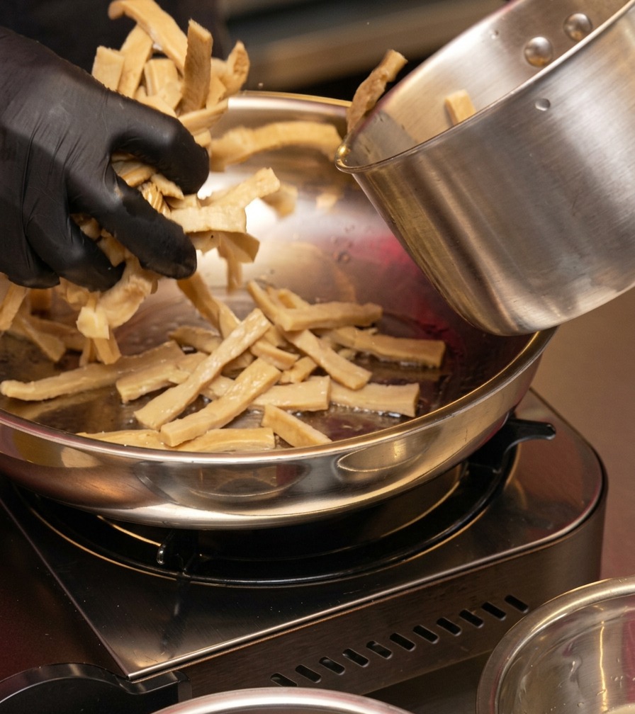 A person in black gloves adding a handful of sliced bamboo shoots from a metal pot into the hot frying pan.