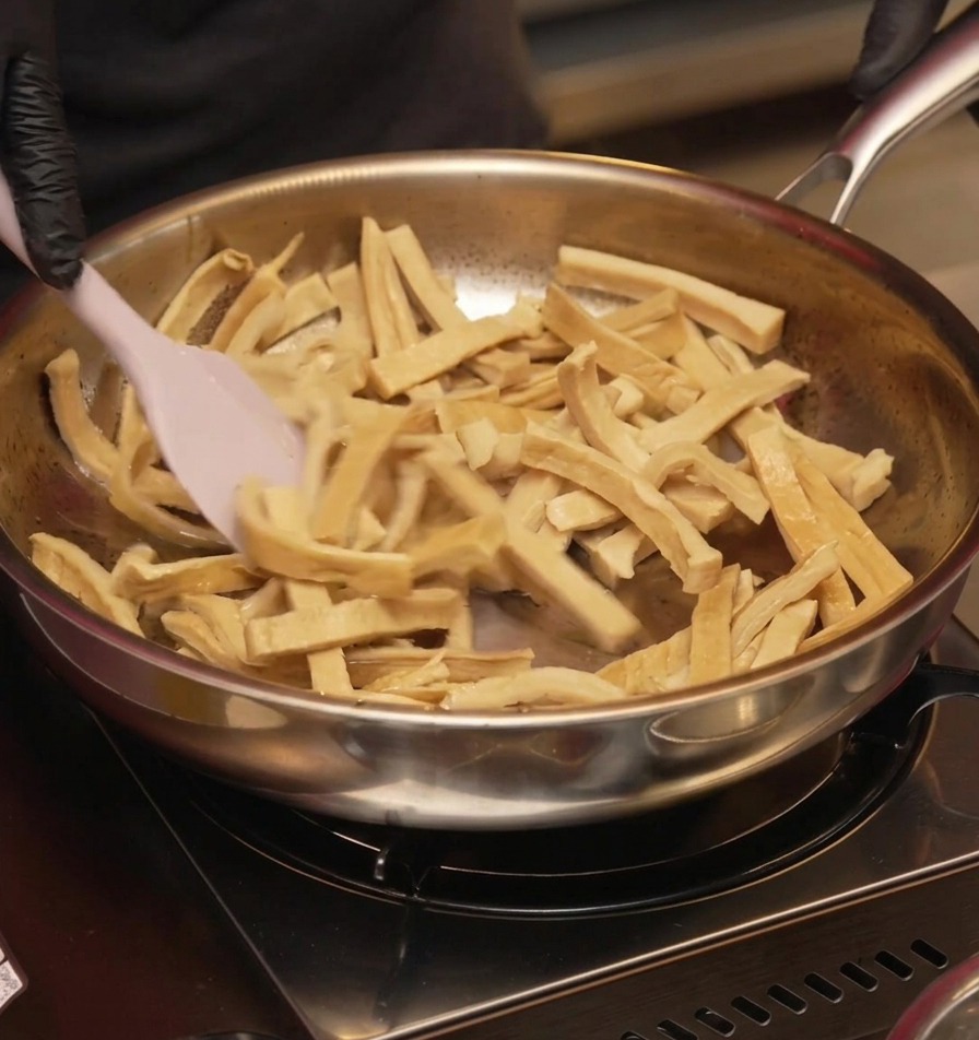 Sliced bamboo shoots being stir-fried in a pan with a pink silicone spatula until they appear dry and lightly browned.