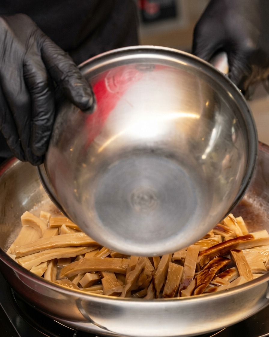 A stream of dark soy sauce being poured from a metal bowl into the pan containing bamboo shoots and liquid.