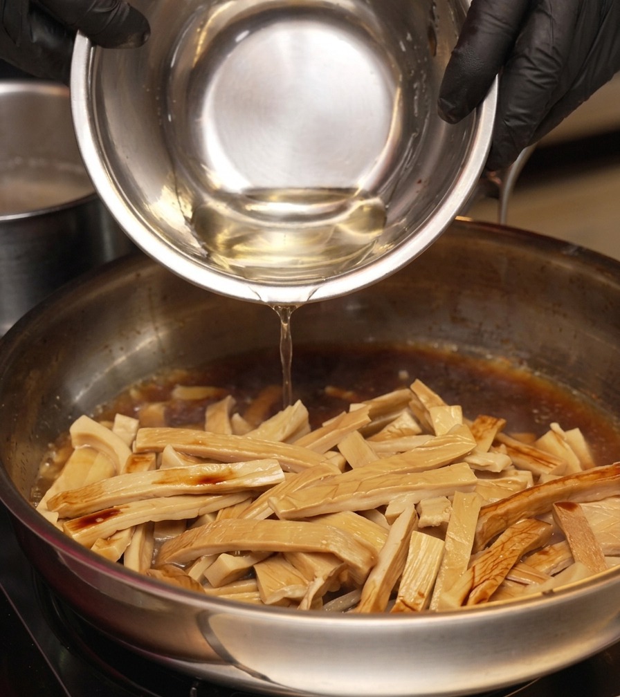 A person wearing black gloves pouring a clear liquid from a metal bowl into a skillet containing bamboo shoots and a dark braising sauce.
