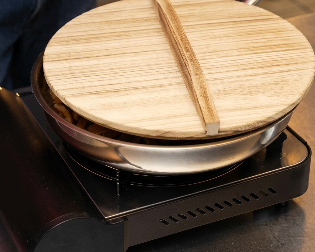 A stainless steel pan of bamboo shoots on a portable gas stove being covered with a traditional wooden lid.