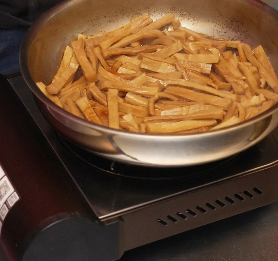 Sesame oil being drizzled into a pan of seasoned bamboo shoots that are stir-frying in a reduced, dark sauce.