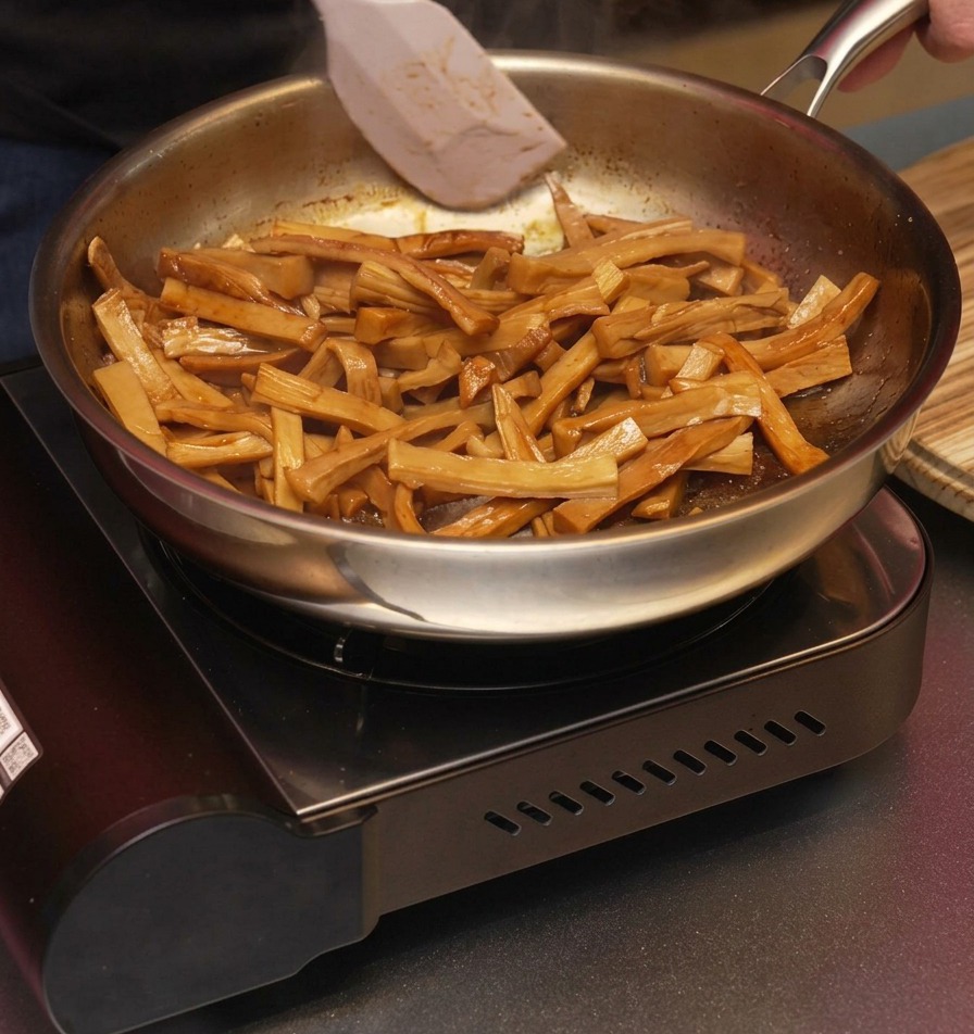 A white spatula tossing glazed, brown bamboo shoots in a frying pan to ensure they are evenly coated in the aromatic sesame oil.