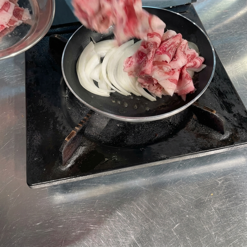 Thinly sliced raw beef rolls being placed into a pan over sautéed white onions.