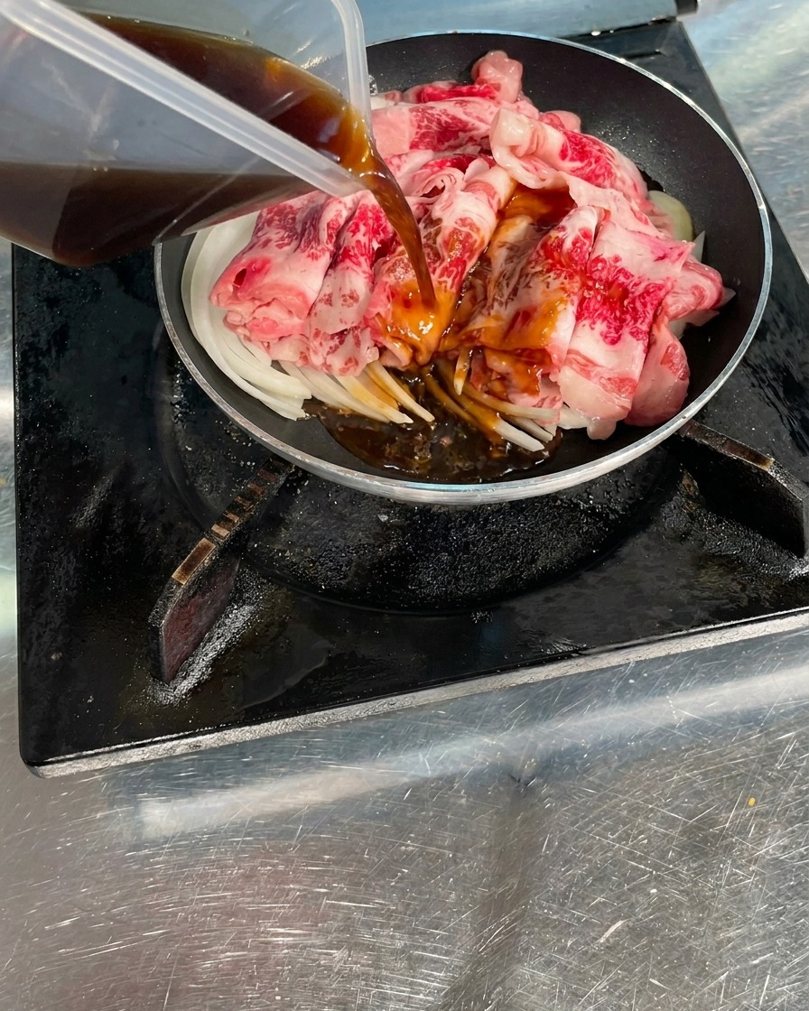 Dark brown savory sauce being poured from a clear container over raw beef slices and onions in a cooking pan.