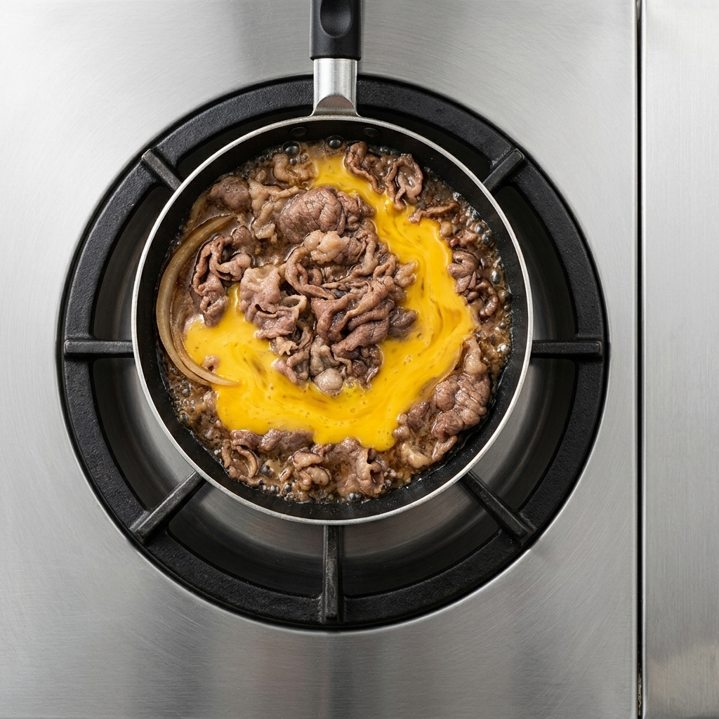 Yellow beaten egg liquid spread over simmered beef slices and onions inside a flat-bottomed pan.