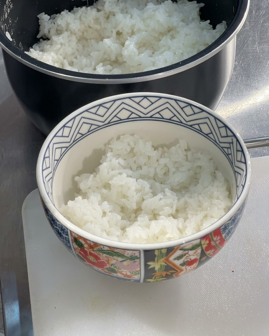 A patterned ceramic bowl half-filled with freshly steamed, fluffy white rice sitting on a cutting board next to a rice cooker.