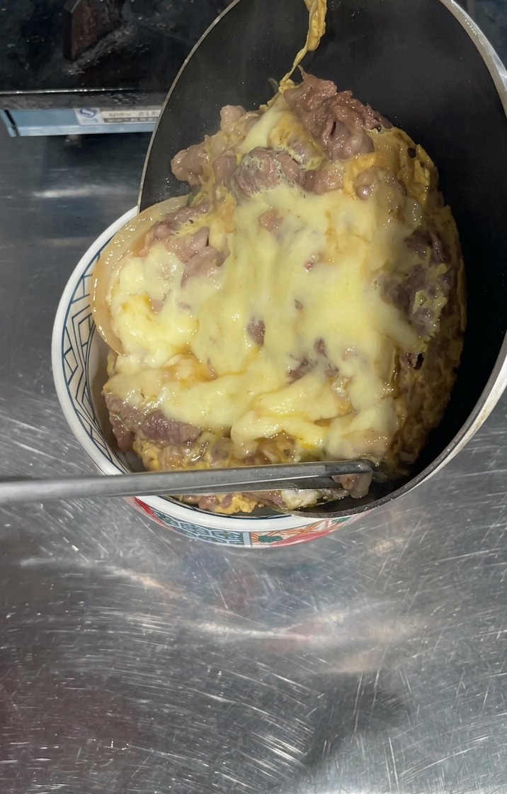 A pan being tilted over a bowl of white rice, with a thick layer of cooked beef and melted yellow cheese sliding out to cover the surface.