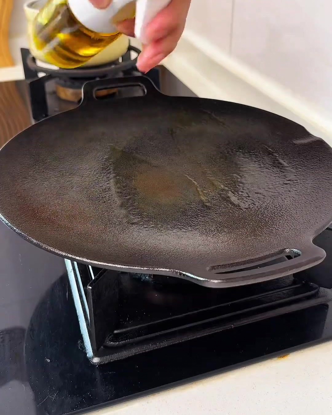 A hand spraying a fine mist of golden cooking oil onto a clean, black circular griddle over a stovetop.
