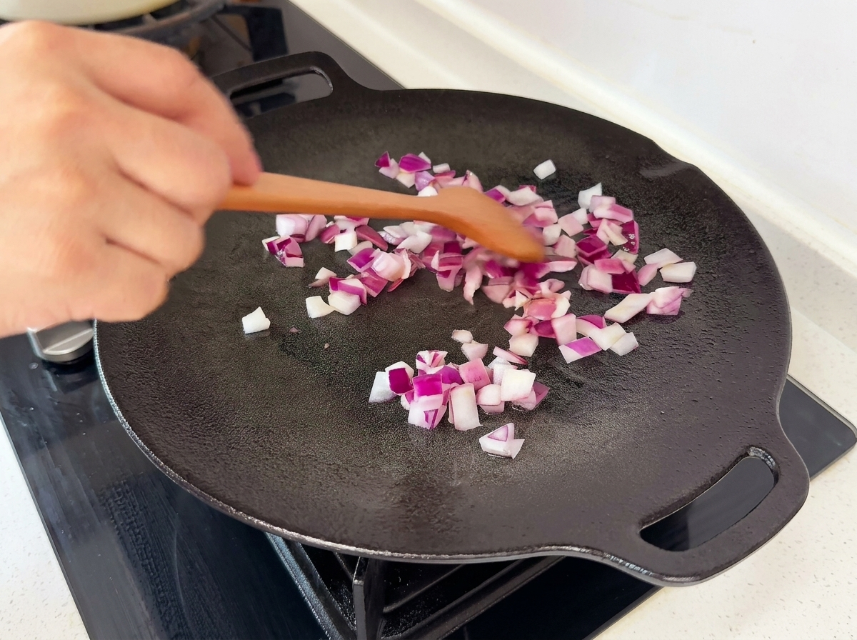 Diced red onions being tossed on a black flat pan with a wooden spatula.